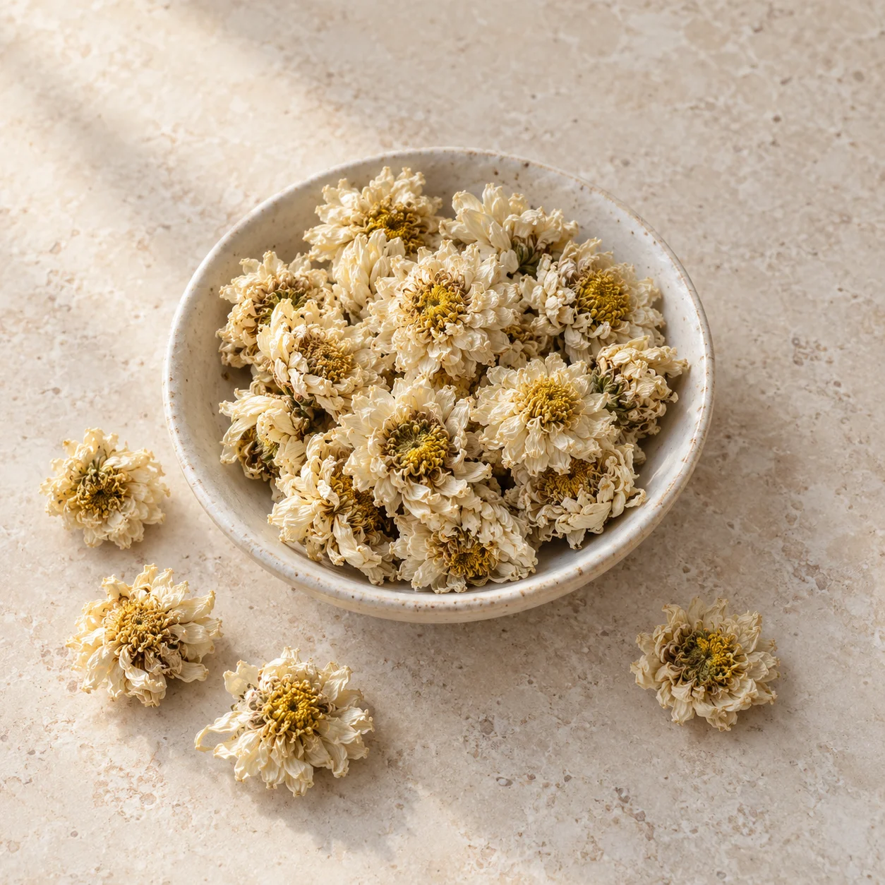 Dried white culinary chrysanthemum flower heads in a shallow ceramic dish on pale stone; editorial illustration, not supplier photography